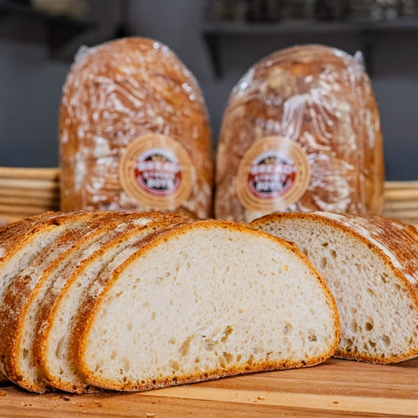 Sliced artisan sourdough bread on a wooden board with two wrapped loaves in the background.