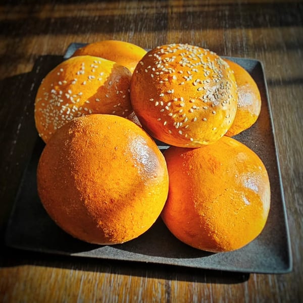 A black plate holds five golden-brown burger buns, some topped with sesame seeds, on a wooden table.
